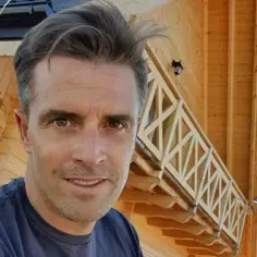 Staszek Karpiel-Bułecka uśmiecha się przed drewnianym domem z charakterystyczną balustradą.