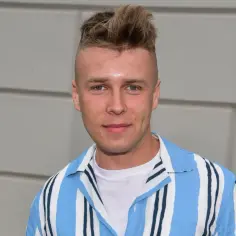 Młody mężczyzna z charakterystyczną fryzurą, ubrany w białą koszulkę i niebiesko-białą koszulę w paski. Czy to właśnie tak ma na imię Skolim?