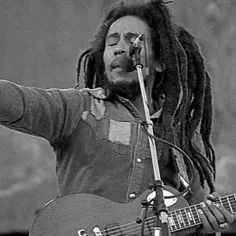 Bob Marley, z charakterystycznymi dredami, śpiewa do mikrofonu, grając na gitarze. Jego muzyka inspirowała wielu, w tym jego liczne bob marley dzieci.