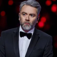 Kuba Badach w czarnym smokingu z muszką, z siwiejącymi włosami i brodą, na tle czerwonych świateł.