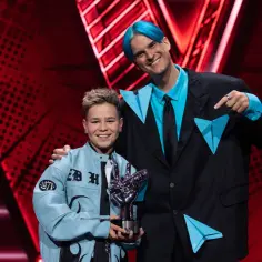 Chłopiec z błękitną kurtką i trofeum w kształcie dłoni, obok niego mężczyzna z niebieskimi włosami. To on wygrał The Voice Kids!