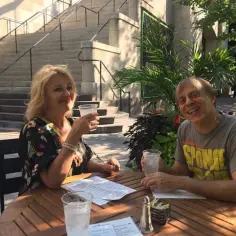 Tom Logan, mąż Majki Jeżowskiej, uśmiecha się do zdjęcia, siedząc przy stoliku na zewnątrz z drinkiem.