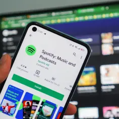 Dłoń trzyma smartfon z otwartą aplikacją Spotify. W tle widać ekran komputera z ikonami aplikacji.