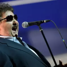 Krzysztof Krawczyk wiek nie przeszkadza mu w śpiewaniu z pasją. Wokalista w okularach przeciwsłonecznych i garniturze przed mikrofonem.