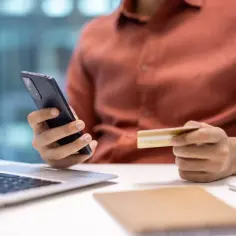 Osoba trzymająca telefon i kartę kredytową przy laptopie.