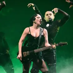 Lady Gaga koncert, w czarnym stroju z gitarą, śpiewa na scenie otoczona tancerzami w podobnych stylizacjach.