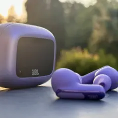 Fioletowe słuchawki bezprzewodowe JBL i ich etui na tle natury.