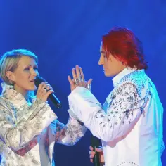 Ich troje na Eurowizji: wokalistka z mikrofonem i mężczyzna z rudymi włosami, oboje w ozdobnych strojach, na niebieskim tle.