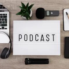 Laptop, słuchawki, mikrofon, dyktafon, notatnik i telefon leżą obok tablicy z napisem PODCAST.
