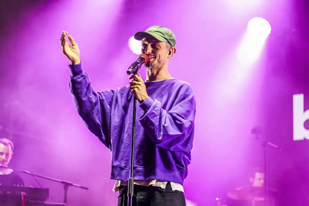 Vito Bambino w fioletowej bluzie i zielonej czapce śpiewa na scenie, podnosząc rękę. Jego wzrost jest imponujący.