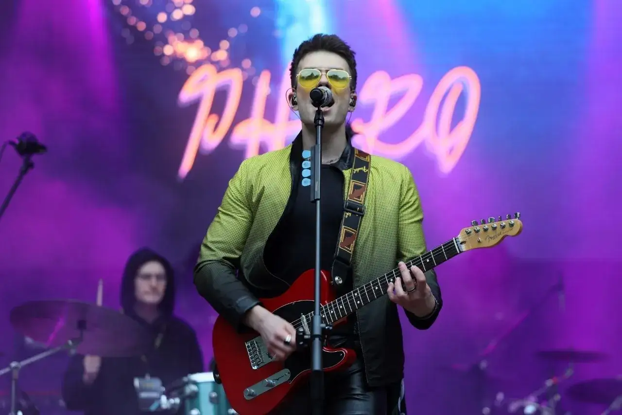 Kris Cugowski w żółtych okularach i zielonej kurtce gra na czerwonej gitarze na tle fioletowej sceny.