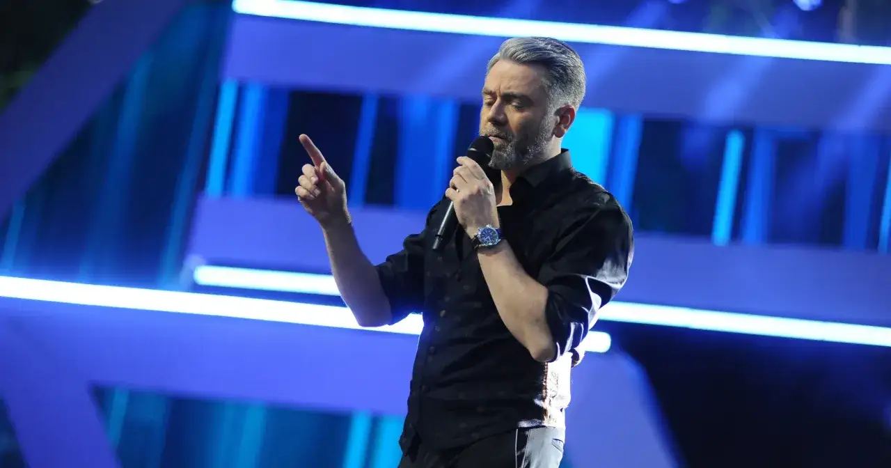 Kuba Badach, w czarnej koszuli, z mikrofonem w dłoni, gestykuluje na tle niebieskich świateł.