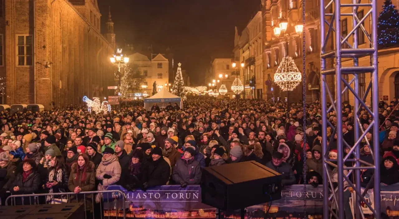 Tłum ludzi na rynku w Toruniu, świętujący Sylwestra z Polsatem. W tle świąteczne iluminacje i choinka.