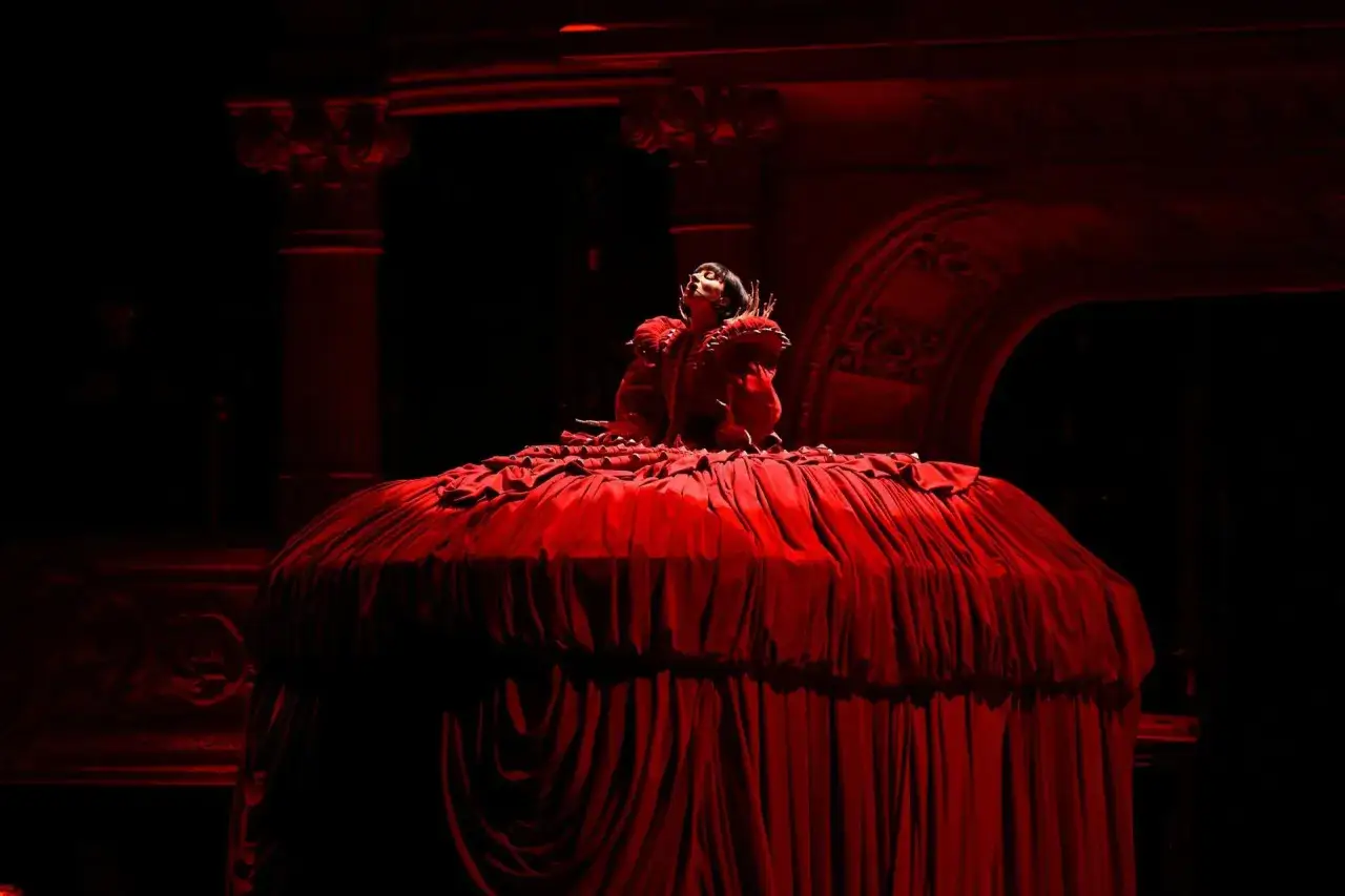 Lady Gaga w czerwonej, obszernej sukni na scenie podczas koncertu. Artystka wznosi głowę ku górze, otoczona teatralnym mrokiem.