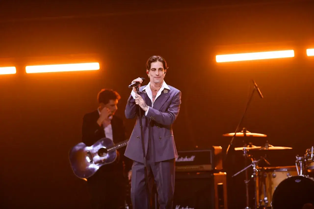 Damiano David na koncercie, w eleganckim garniturze, śpiewa do mikrofonu. W tle gitarzysta i perkusja.