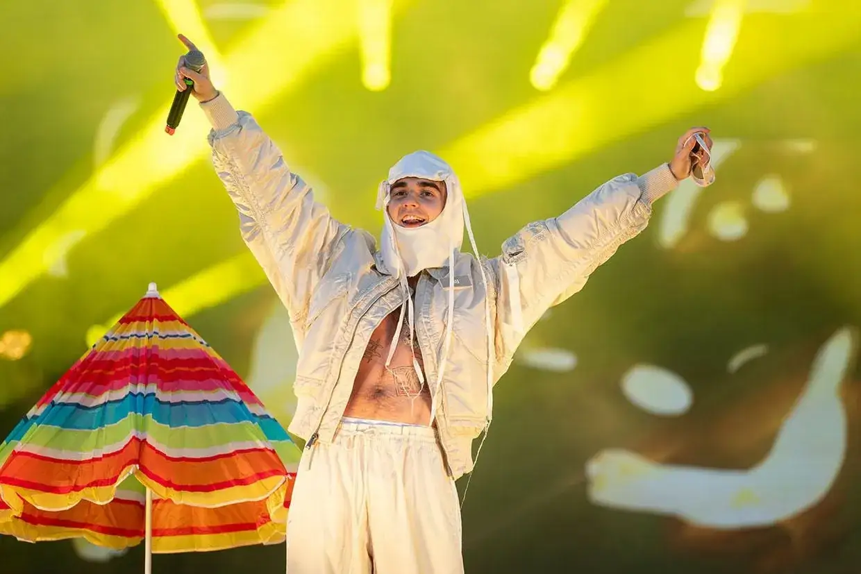 Artysta w beżowej kurtce i czapce z kominem, z mikrofonem w dłoni, świętuje na scenie. Obok kolorowy parasol. Ile mata ma lat?